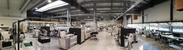 Photo grand angle de l'usine de production avec les lignes d'assemblage, les différentes machines et postes de travail. 
L'usine est spacieuse et très lumineuse.